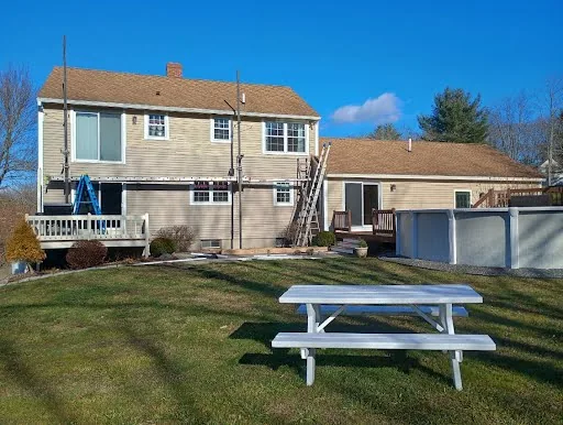 Rear yard view of a beige two-story home during exterior siding work with ladders in place
